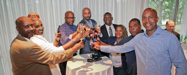 Eight smiling alumni standing around a small round table raise their drinks in a toast.